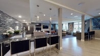 This single-floor amenity space is designed with a contemporary aesthetic, combining functionality and sophisticated style throughout its open layout. The wet bar area serves as a central focal point, featuring dark granite countertops, sleek black bar stools with chrome frames, and a striking mosaic tile accent wall in charcoal and black tones. Pendant lighting adds an intimate ambiance above the bar counter.

Adjacent to the bar, a dedicated media lounge showcases a large flat-screen television mounted on a dark wood feature wall, flanked by additional screens and complemented by orange leather ottomans atop a graphic wave-patterned area rug. This zone transitions seamlessly into a comfortable lounge area furnished with neutral-toned sofas and accent chairs, accented with decorative throw pillows and curated artwork.

Light wood-style flooring anchors the open circulation areas, providing a visual contrast to the darker lounge zones. Large floor-to-ceiling windows and a glass sliding door flood the space with natural light and offer direct views of and access to an outdoor terrace. A glass-enclosed conference or business room is accessible via double doors, adding a practical element to the overall layout. Recessed lighting throughout ensures a well-illuminated, welcoming environment.