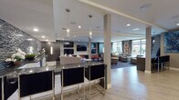 This single-floor amenity space is designed with a contemporary aesthetic, combining functionality and sophisticated style throughout its open layout. The wet bar area serves as a central focal point, featuring dark granite countertops, sleek black bar stools with chrome frames, and a striking mosaic tile accent wall in charcoal and black tones. Pendant lighting adds an intimate ambiance above the bar counter.

Adjacent to the bar, a dedicated media lounge showcases a large flat-screen television mounted on a dark wood feature wall, flanked by additional screens and complemented by orange leather ottomans atop a graphic wave-patterned area rug. This zone transitions seamlessly into a comfortable lounge area furnished with neutral-toned sofas and accent chairs, accented with decorative throw pillows and curated artwork.

Light wood-style flooring anchors the open circulation areas, providing a visual contrast to the darker lounge zones. Large floor-to-ceiling windows and a glass sliding door flood the space with natural light and offer direct views of and access to an outdoor terrace. A glass-enclosed conference or business room is accessible via double doors, adding a practical element to the overall layout. Recessed lighting throughout ensures a well-illuminated, welcoming environment.
