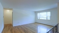 This single-floor space presents a clean and straightforward design, characterized by its minimalist aesthetic and well-maintained finishes. The interior features warm-toned hardwood flooring that runs throughout, adding a natural richness to the otherwise neutral palette of crisp white walls and ceilings. A large window allows generous natural light to pour into the main living area, creating a bright and open atmosphere. Beneath the window, a baseboard heating unit provides climate comfort throughout the colder months. The walls are smooth and freshly finished, offering a blank canvas ready for personalization. The layout is open and unobstructed, providing a flexible use of space suitable for a variety of configurations. Electrical outlets are conveniently positioned along the walls, supporting practical everyday use. The overall ambiance is calm and understated, with the combination of light wood floors and white surfaces giving the space a clean, airy feel. This space is well-suited for those who appreciate simplicity and functionality in their environment.