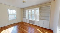 This single-floor space showcases a thoughtfully designed interior that blends classic architectural details with functional built-in storage. The main living area is anchored by a wall of white built-in shelving and cabinetry flanking a trio of double-hung windows, providing both generous storage and a charming window seat arrangement. The built-ins extend from floor to ceiling, offering open shelving above and closed cabinet storage below, making the space as practical as it is visually appealing. A traditional radiator beneath the central windows hints at the space's classic character. Rich, warm-toned hardwood floors run throughout, polished to a high sheen that reflects the natural light streaming in from multiple windows on adjacent walls. The cream-toned walls and white trim create a clean, airy aesthetic that complements the warmth of the wood flooring. A flush-mount ceiling fixture with a brass finish adds a subtle traditional touch overhead. The overall ambiance is bright, open, and inviting, with classic architectural elements that lend the space a timeless, well-crafted character.