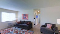 Located at 124 Carolina Ave in Atlantic City, New Jersey, this single-floor residence offers a practical and welcoming layout designed for comfortable everyday living. The living area serves as the heart of the home, featuring tile flooring, smooth ceilings, and neutral wall tones that create a clean, airy backdrop. A large area rug with a bold, multicolored circular pattern anchors the seating arrangement, which includes a charcoal sofa and matching accent chair. Vibrant abstract artwork adorns the walls, adding pops of color and personality to the space. Natural light filters in through a window on the adjacent wall, brightening the room throughout the day. A central hallway connects the living area to the rest of the unit, providing a clear sightline to the bedroom and additional rooms beyond. The bedroom is visible at the end of the hall, hinting at a generous and well-organized private space. Flush ceiling lighting in the hallway ensures consistent illumination throughout. The overall ambiance is casual yet curated, blending functional design with cheerful, expressive décor touches that give the space a lively and inviting character throughout.