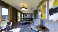 This sophisticated single-floor amenity lounge is thoughtfully designed with a cohesive contemporary aesthetic that blends bold color with refined furnishings. The space is defined by a striking color palette of charcoal grey, muted blues, and vibrant yellow accents — carried through the patterned carpeting, upholstered seating, and decorative wall details adorned with yellow stud trim. Large windows dressed in chartreuse drapes flood the space with natural light, creating a bright and inviting atmosphere. A plush blue wingback chair and round gold side table anchor the lounge area, complemented by an oversized abstract artwork in yellow and grey tones. The dining area features a dark round table surrounded by upholstered chairs, positioned beneath dramatic drum pendant lighting that adds a modern, layered illumination effect. Toward the rear, a wet bar area with mosaic tile backsplash, pendant lights, and dark cabinetry provides a functional yet stylish entertaining hub. The patterned carpet throughout ties the design elements together, reinforcing the space's intentional and polished character. Overall, this lounge offers a welcoming, design-forward environment well-suited for both social gatherings and relaxed everyday use.