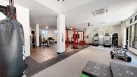 This spacious fitness center is designed to support a comprehensive range of workout disciplines within a single, open floor plan. The interior is defined by crisp white walls, structural columns, and rubber-tiled flooring throughout, providing both a clean aesthetic and a practical, durable surface for heavy equipment use. Recessed and track lighting supplement the abundant natural light that streams in through large, floor-to-ceiling windows, offering views of outdoor greenery and maintaining a bright, energizing atmosphere.

The strength training area is well-appointed with selectorized machines, a leg press, free-weight benches, and an extensive dumbbell rack backed by large mirrors that enhance the sense of space. A bold red power rack serves as a central functional training station, accompanied by TRX suspension straps, a barbell, and weight plates. The functional fitness zone includes plyo boxes, a heavy punching bag, a tire, and battle rope anchors, providing versatile options for high-intensity training. Cardio equipment, including stair climbers and treadmills, is positioned near the windows to maximize natural light exposure. A staircase with metal railings adds architectural character, and ceiling-mounted HVAC units ensure a comfortable environment throughout.