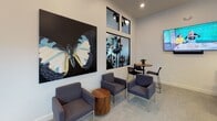 This thoughtfully designed single-floor space blends contemporary aesthetics with functional versatility. The main lounge area makes a striking first impression with high ceilings, a large-scale butterfly canvas artwork, and an adjacent palm tree photographic mural framed by tall windows that invite natural light. Comfortable grey lounge chairs and a round wooden side table create a relaxed seating arrangement, while a wall-mounted display screen adds a modern touch. A casual high-top table and bistro chairs near the windows offer an alternative spot for informal gatherings. A dedicated dining nook features built-in banquette seating with patterned upholstery, white tables, and statement pendant lighting, flanked by a pair of expressive abstract artworks in muted blue and grey tones that lend the area an artistic ambiance. The conference room is a standout feature, anchored by a long wooden table surrounded by sleek wire-frame chairs. Bold black-and-white patterned floor-to-ceiling curtains frame a large window overlooking greenery, while a dramatic multi-bulb chandelier and tall potted plants complete the sophisticated setting. Throughout the space, light grey carpeting, crisp white walls, and carefully selected décor create a cohesive, modern atmosphere ideal for both collaboration and relaxation.