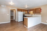 This single-story unit is designed with a practical, open layout that connects the kitchen and living areas seamlessly. The kitchen is well-equipped with warm-toned wood cabinetry offering generous storage, light-colored countertops, a black electric range, a white refrigerator, and a double-basin sink. A peninsula counter separates the kitchen from the living space, providing additional prep area and a natural divider between the two zones. The living area features durable wood-look flooring throughout and is bathed in natural light from a large sliding glass door fitted with vertical blinds, which also offers views of the surrounding greenery. A standard entry door provides convenient access to the exterior. The bedroom is carpeted for added comfort and includes a sizable window with vertical blinds that allow for adjustable natural light. The bathroom contains a tiled shower-tub combination, a vanity with a dark granite-style countertop, wood-look flooring, and a ceiling exhaust vent. Textured ceilings and warm tan-painted walls are consistent throughout, contributing to a cohesive, functional interior aesthetic.