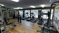 This fitness center is designed to support a well-rounded exercise routine within a single-level layout. The space features hardwood-style flooring throughout, providing a clean and durable surface suited for high-traffic use. Large windows line the walls, allowing generous amounts of natural light to fill the room and offering views of the surrounding outdoor greenery, contributing to an open and energizing atmosphere. The dark earth-tone walls provide a grounding contrast to the bright, naturally lit interior.

The room is equipped with multiple treadmills, elliptical trainers, and additional cardio machines, each fitted with individual TV monitors for entertainment during workouts. Protective rubber mats are placed beneath the equipment to safeguard the flooring. A full-length mirror runs along one wall, visually expanding the space and allowing users to monitor their form. Overhead fluorescent lighting supplements the natural light, ensuring the room remains well-lit at all hours. A water cooler is also visible, adding a convenient amenity for gym users. Overall, the fitness center balances functionality and comfort effectively.