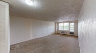 This single-floor unit is designed with a practical, straightforward layout. The living area is generously proportioned, featuring wall-to-wall beige carpeting and a textured ceiling. A wall-mounted air conditioning unit serves the space, and a wide window with an adjacent door provides natural light and outdoor views, creating a bright and open atmosphere.

The kitchen is efficiently arranged with warm wood-toned cabinetry extending to the ceiling for ample storage. White laminate countertops provide a clean contrast, and the space is equipped with a stainless steel single-basin sink, a white refrigerator, a gas range, and a dishwasher. Light tile flooring and a window with slatted blinds complete the functional cooking area, which also features a baseboard heater.

The bedroom is notably spacious, also finished with beige carpet and a textured ceiling. Two sets of bifold closet doors offer generous built-in storage side by side.

The bathroom is finished with white cabinetry, a vanity with a mirror and lighted fixture above, and mosaic tile flooring accented with subtle blue dot tilework on the walls. Additional open shelving provides practical storage, contributing to the room's organized, clean aesthetic.