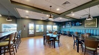 Located in Bend, Oregon, this pub-style dining space offers a well-appointed, single-floor layout designed for comfortable communal gathering and dining. The room features warm light hardwood flooring throughout, complemented by rich teal and olive-green painted walls that create a casual yet inviting atmosphere reminiscent of a classic neighborhood pub.

The space is anchored by a central wraparound bar with dark countertops and cushioned bar-height seating, backed by a functional service area with overhead cabinetry and prep surfaces. Industrial-style pendant lights hang from a drop ceiling with recessed lighting, adding character and focused illumination throughout the room.

Dining seating is generously distributed across the floor, with a mix of freestanding tables paired with upholstered chairs and striped booth seating along one wall. Decorative pub-themed signage and framed artwork adorn the walls, reinforcing the establishment's relaxed, social character.

Multiple windows along the exterior walls allow natural light to filter in, offering views of the surrounding outdoor area. A service window on one wall provides a direct connection to an adjacent area. The open floor plan ensures ease of movement between the bar and dining sections, making this space well-suited for both casual dining and social gatherings.