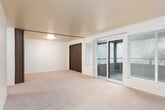 This single-floor residence is thoughtfully laid out to maximize comfort and functionality. The living area is generously proportioned, featuring soft wall-to-wall carpeting, neutral cream-toned walls, and multiple windows fitted with horizontal blinds that allow for controlled natural light. A sliding glass door opens directly onto a private balcony, offering pleasant outdoor views of surrounding greenery and providing a seamless indoor-outdoor connection.

The galley-style kitchen is efficiently designed with rich warm-toned wood cabinetry lining both sides, offering ample upper and lower storage. White appliances — including a refrigerator, electric range, and dishwasher — complement the light countertops and tile-style flooring, creating a clean, practical workspace. A double-basin sink is positioned conveniently along one wall.

An additional carpeted room, accessible via a dark wood-paneled door, provides a versatile space that could serve as a bedroom or flexible living area. It features its own window with blinds and access to a sliding door.

The bathroom is finished with tile flooring, a wide vanity counter with an integrated sink, warm wood cabinetry below, and a large wall mirror that enhances the sense of space. The overall ambiance throughout is clean, neutral, and move-in ready.