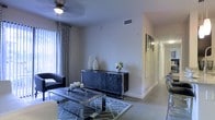 This single-story space presents a thoughtfully designed living area that combines modern aesthetics with functional comfort. The living room is anchored by a geometric-patterned area rug and a sleek, dark-toned coffee table with metallic accents, complemented by a matching dark wood credenza that serves as a striking focal point along the main wall. Decorative vases and a fresh floral arrangement atop the credenza add an elegant finishing touch.

Natural light filters into the space through a large window dressed with patterned curtains, while a ceiling fan overhead ensures comfortable airflow throughout. The light-toned tile flooring runs consistently through the space, enhancing the sense of openness. Neutral-painted walls provide a versatile backdrop that allows the darker furnishings to stand out effectively.

A hallway visible from the living area leads further into the unit, with a bold black-and-white artwork piece adorning the wall, adding a contemporary artistic element. The overall ambiance is modern and polished, with a carefully curated mix of textures and tones that give the space a sophisticated, welcoming character.
