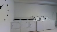 This shared laundry facility is a clean, single-floor space purpose-built to accommodate multiple users efficiently. The room features a row of commercial-grade front-load dryers paired with top-load washing machines, all finished in white and equipped with digital control panels for ease of use. The machines are arranged in a neat, organized line along the wall, maximizing floor space and allowing for comfortable movement throughout the room. The interior is characterized by bright white walls and a light-toned floor, contributing to a clean and functional atmosphere. Overhead lighting ensures the space is well-lit, enhancing visibility and usability. Water hookups are visibly integrated into the installation, indicating permanent plumbing infrastructure supporting the machines. The minimalist design of the room keeps the focus on utility, with no unnecessary furnishings or clutter. The layout is straightforward and accessible, making laundry tasks straightforward for all users. Overall, the space conveys a practical, no-frills environment that prioritizes cleanliness, organization, and operational efficiency for communal laundry needs.