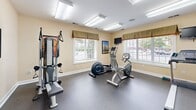 Located at 901 Forest Lake Ct in Newport News, Virginia, this dedicated exercise room offers a well-equipped fitness facility designed to support a well-rounded workout routine. The space features a durable speckled rubber floor, ideal for supporting heavy equipment and providing comfort underfoot during exercise sessions.

The room is outfitted with a variety of fitness equipment, including a treadmill, elliptical trainer, recumbent stationary bike, and a multi-function cable strength training machine with an adjustable bench. A full-length mirror spans one wall, amplifying the sense of space and allowing users to monitor their form during workouts.

Bright, recessed fluorescent panel lighting illuminates the room evenly from the ceiling, while multiple windows dressed with valances and horizontal blinds allow natural light to filter in and provide views of the surrounding outdoor area. A wall-mounted flat-screen television is positioned for easy viewing during cardio sessions, adding an element of entertainment to workouts.

The walls are finished in a warm, neutral cream tone, contributing to a clean and inviting atmosphere. A thermostat on the wall ensures climate control for year-round comfort. The room's efficient, purpose-driven layout makes it a practical and functional fitness space for daily use.