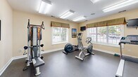 Located at 901 Forest Lake Ct in Newport News, Virginia, this dedicated exercise room offers a well-equipped fitness facility designed to support a well-rounded workout routine. The space features a durable speckled rubber floor, ideal for supporting heavy equipment and providing comfort underfoot during exercise sessions.

The room is outfitted with a variety of fitness equipment, including a treadmill, elliptical trainer, recumbent stationary bike, and a multi-function cable strength training machine with an adjustable bench. A full-length mirror spans one wall, amplifying the sense of space and allowing users to monitor their form during workouts.

Bright, recessed fluorescent panel lighting illuminates the room evenly from the ceiling, while multiple windows dressed with valances and horizontal blinds allow natural light to filter in and provide views of the surrounding outdoor area. A wall-mounted flat-screen television is positioned for easy viewing during cardio sessions, adding an element of entertainment to workouts.

The walls are finished in a warm, neutral cream tone, contributing to a clean and inviting atmosphere. A thermostat on the wall ensures climate control for year-round comfort. The room's efficient, purpose-driven layout makes it a practical and functional fitness space for daily use.