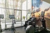 Located at 1410 Fifth Street in Santa Monica, California, this contemporary fitness facility is thoughtfully designed to support a full range of training modalities. The space features soaring double-height ceilings in the main gym area, with expansive clerestory windows along the upper walls that flood the floor with natural light and offer glimpses of lush greenery outside.

The main workout floor is finished with durable, speckled rubber flooring and houses an impressive array of free weights, including a full rack of dumbbells, a Precor cable machine, and resistance training accessories such as resistance bands and a speed bag. A glass-railed staircase adds a sleek architectural element while connecting the upper observation level, where a wall-mounted TV overlooks the training floor below.

A dedicated cardio zone features a stair climber, elliptical, and treadmill arranged in front of a bold, floor-to-ceiling panoramic wall mural depicting an open road at sunset, creating an immersive and motivating environment. A separate functional training area with blue rubber flooring includes a multi-purpose rig and battle ropes, framed by a translucent skylight ceiling and bamboo garden views. Built-in shelving provides organized storage throughout. Private rooms along one side of the space offer additional flexibility for focused training or consultations.
