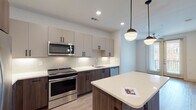 This single-floor residence showcases a thoughtfully designed open-concept layout with modern finishes throughout. The kitchen serves as a central highlight, featuring a two-tone cabinet design that pairs warm wood-grain lower cabinets with light greige upper cabinets, all accented by sleek matte black hardware. A full suite of stainless steel appliances includes a French door refrigerator, electric range, over-the-range microwave, and dishwasher. A subway tile backsplash adds texture, while under-cabinet lighting illuminates the clean white quartz countertops. A freestanding center island provides additional workspace and casual seating potential, topped with a matching quartz surface and pendant globe lights suspended above. Light wood-tone luxury vinyl plank flooring runs through the main living and kitchen areas, contributing to the bright, airy atmosphere created by recessed lighting throughout. The bedroom features plush neutral carpet and crisp white walls, offering a calm retreat. The bathroom is finished with large-format tile flooring, a soaking tub with subway tile surround, a white quartz vanity with wood cabinetry, and direct access to a spacious walk-in closet equipped with built-in shelving and hanging rods.
