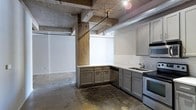 This single-floor space showcases a compelling blend of raw industrial character and modern functionality. The open layout is defined by exposed concrete ceilings with visible ductwork and conduit running overhead, supported by original concrete and wood columns that serve as striking architectural focal points. Polished concrete floors extend throughout, reinforcing the industrial aesthetic while adding durability.

The kitchen is well-appointed with gray shaker-style cabinetry arranged in an L-shaped configuration, offering generous storage and counter space. White subway tile backsplash provides a clean contrast against the cabinetry, while stainless steel appliances — including a gas range and built-in microwave — add a modern, functional edge. A stainless steel sink with a contemporary faucet completes the setup.

The adjacent open area benefits from bright white walls that reflect light effectively, creating a sharp contrast with the rugged structural elements. Track lighting mounted to the ceiling provides targeted illumination throughout the space. The overall ambiance balances utilitarian, loft-style character with practical, updated finishes suited for a variety of uses.