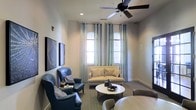 This single-floor space presents a thoughtfully designed living area that balances comfort and style. The room features two arched windows dressed with sheer, patterned curtains that allow natural light to filter softly throughout the space, enhancing the neutral gray-toned walls. A ceiling fan with dark-finished blades is centrally mounted, offering both practical airflow and a subtle design element overhead. The flooring transitions between carpet in the seating area and hard surface near the dining space, subtly delineating functional zones. The living area is anchored by a beige loveseat paired with two bold blue leather wingback chairs, creating a balanced and inviting conversation area around a round white-topped coffee table with a geometric metal base. Wall art in muted tones adds visual interest without overwhelming the space. Adjacent to the living area, a glimpse of a dining setup is visible, suggesting a seamless open-concept flow. The overall ambiance is calm and contemporary, with a cohesive mix of soft textiles, neutral tones, and carefully chosen furnishings that create a welcoming and functional environment.