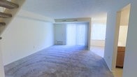 This two-floor space presents a straightforward, functional layout with a focus on practicality and openness. The main living area is finished with wall-to-wall carpeting in a neutral tone, providing a soft underfoot feel that extends throughout the room. Large windows fitted with vertical blinds allow for adjustable natural light, brightening the interior and creating an airy atmosphere. The walls are painted in a clean, light neutral palette, contributing to the overall sense of openness. A textured ceiling adds subtle visual interest overhead, while the room's generous proportions offer ample space for furniture arrangement. Baseboard vents are visible along the lower walls, indicating a central climate control system. A secondary windowed area is partially visible to the side, suggesting additional rooms that continue the same design language of light tones and carpeted flooring. The overall ambiance is clean and understated, providing a blank canvas that allows for personalization. Spanning two floors, the space offers a layered living arrangement suitable for a variety of lifestyle needs.