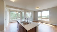 Located on the fourth floor at 6979 Birdseye Avenue NE in Lacey, Washington, Unit 408 is a thoughtfully designed residence that blends modern finishes with comfortable, functional living. The open-concept living and dining area features warm hardwood-style flooring and an abundance of natural light streaming through multiple large windows, offering sweeping views of the surrounding tree line and neighborhood rooftops. The kitchen is anchored by a central island with a double-basin sink and crisp white countertops, complemented by rich dark cabinetry, black appliances including a refrigerator, electric range, and over-the-range microwave, and a convenient in-unit stacked washer and dryer closet nearby. The bedrooms are carpeted for comfort, with neutral-toned walls and generous windows — one bedroom features a sliding glass door opening to a private balcony overlooking lush evergreen trees. The bathrooms are finished with wood-look flooring, dark vanity cabinetry, white countertops, and a full tub/shower surround. Throughout the unit, a consistent palette of warm neutrals and clean white trim creates a bright, cohesive atmosphere well-suited for everyday living.