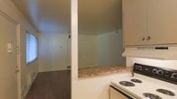 This single-floor space offers a functional and straightforward layout designed for comfortable everyday living. The kitchen is equipped with an electric coil-burner range and features white cabinetry overhead, providing ample storage. A defining characteristic of the kitchen is the pass-through counter that opens directly into the adjoining living area, creating a connected, open feel between the two spaces. This design element encourages interaction and allows natural sightlines across the main living zone. The countertop surfaces display a warm, neutral tone that complements the light-colored walls found throughout the space. The living area is generously proportioned, with smooth walls painted in a soft, neutral hue and ceilings that lend an open, airy quality to the room. A white interior door provides access to additional areas of the home, contributing to the clean, minimal aesthetic. The flooring throughout the main areas consists of dark hardwood-style planks, offering both durability and a grounded visual contrast to the light walls and cabinetry. Ceiling vents indicate a central HVAC system for year-round climate comfort. Overall, the space presents a clean, no-frills interior well-suited for those seeking a practical and move-in-ready environment.