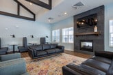 Located at 6207 Harvey St in Norton Shores, Michigan, this single-floor community clubhouse is designed to offer a well-appointed, versatile gathering space. The architecture is defined by dramatic vaulted ceilings accented with bold dark wood beam detailing, creating an open and airy atmosphere throughout the main lounge area. Wood-look flooring flows consistently across the space, tying together distinct zones with a warm, cohesive aesthetic.

The main lounge area serves as the centerpiece, featuring a floor-to-ceiling slate tile fireplace with a rustic wood mantle and a wall-mounted television above. Leather sofas arranged over a colorful area rug invite comfortable seating, while a row of upholstered booth-style tables along one wall provides a more intimate dining or work setting with individual task lighting.

An adjacent seating lounge is painted in a rich navy blue accent color and lined with large windows that flood the space with natural light and neighborhood views. Cognac-toned leather armchairs are arranged around round marble-top tables, creating a casual yet refined conversation area.

The kitchen features a large granite-topped island with bar seating, globe pendant lighting, stainless steel appliances, and a striking hexagonal tile backsplash with floating wood shelving, making it ideal for community events and gatherings.