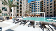 This expansive outdoor amenity area is centered around a resort-style swimming pool, nestled within the interior courtyard of a contemporary multi-story building. The architecture features a neutral sand-toned facade with large windows and private balconies that overlook the pool deck, creating a sense of enclosure while maintaining an open, airy atmosphere under abundant natural sunlight.

The pool itself is generously sized, with clear blue water and clean geometric lines that complement the modern aesthetic. Surrounding the pool is a wide, light-toned tile deck furnished with padded chaise lounges and accent tables, offering ample space for sunbathing and relaxation. Tall palm trees are strategically placed throughout the courtyard, adding a tropical character to the setting.

A white pergola structure anchors the central lounge area, providing shaded seating beneath. Distinctive horizontal wood-slat privacy screens serve as both functional dividers and design elements, adding warmth and texture to the otherwise contemporary palette. Semi-private cabana-style alcoves, lined with these natural wood panels and fitted with cushioned loungers and accent pillows, offer intimate retreats within the larger communal space.

An outdoor grilling and dining area is also present, equipped with stainless steel grills and seating. Raised planters with ornamental grasses and sculptural planters with manicured shrubs contribute greenery throughout the deck, rounding out a well-appointed outdoor living environment.
