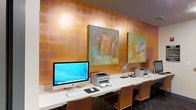 This single-floor business center offers a well-appointed workspace designed to meet professional needs in a polished, aesthetically considered environment. A long, sleek white countertop runs along the length of the room, providing ample workspace for multiple users simultaneously. The stations are equipped with modern all-in-one desktop computers, wireless keyboards, and mice, alongside dedicated printers conveniently positioned for shared use. Networking equipment is also present, ensuring reliable connectivity.

The design aesthetic is elevated by a striking amber-toned feature wall that serves as a warm, luminous backdrop to the workspace. Recessed lighting casts a soft glow across the wall's textured surface, enhancing its rich, burnished appearance. Two large abstract paintings in complementary earth and muted tones are displayed prominently, adding a sophisticated, gallery-like quality to the space. A sculptural starburst accent piece provides an additional decorative touch.

Ergonomic seating in a warm wood-toned finish lines the counter, accommodating multiple users comfortably. The overall ambiance balances functionality with curated interior design, creating a workspace that feels both productive and visually engaging.