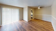 This single-story residence offers a practical and well-organized layout with distinct living areas connected by consistent design elements throughout. The main living area features rich wood-style hardwood flooring, neutral beige walls, and a textured ceiling with a ceiling fan, creating a comfortable and functional space. Large vertical blinds cover a sliding door or window, allowing natural light to filter in. The space opens toward the kitchen through an arched passageway, maintaining a connected flow between rooms.

The galley-style kitchen is efficiently designed with dark cabinetry lining both sides, light countertops, and wood-style flooring that continues from the main living area. It is equipped with a white electric range, refrigerator, and dishwasher, with a window at the far end providing natural light.

The bedroom is finished with plush wall-to-wall carpeting, neutral walls, a ceiling fan with light fixture, and a window fitted with blinds. A walk-in closet offers ample storage space. The textured ceiling is consistent throughout the sleeping area.

The bathroom is clean and straightforward, featuring a full-size soaking tub with white square-tile surround, a showerhead, toilet, and wood-style flooring, completing the functional layout of this well-maintained single-floor residence.