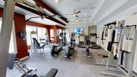 This fitness center offers a comprehensive and well-organized workout environment designed to accommodate a variety of exercise routines. The space is outfitted with an impressive selection of cardio equipment, including multiple treadmills and elliptical machines, all arranged to maximize usability and flow. Strength training needs are met with a selection of weight machines and benches positioned along the perimeter of the room. Durable, rubber-matted flooring provides both safety and comfort underfoot throughout the workout area. The interior design features a striking combination of deep red accent walls and crisp white tray ceilings, giving the space a bold yet polished aesthetic. Two ceiling fans are mounted overhead, ensuring consistent air circulation during intense workouts. Recessed lighting complements the natural light that pours in through large windows along one wall, creating a bright and energizing atmosphere. A wall-mounted flat-screen television is centrally positioned, allowing exercisers to stay entertained while training. The overall layout is thoughtfully planned, offering ample space between equipment for safe and comfortable use, making this fitness center a functional and inviting destination for daily exercise.