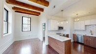 Located at 50 S Jefferson St in downtown Dayton, Ohio, this 2-bedroom, 1-bathroom unit blends historic character with modern finishes. Exposed rustic wood ceiling beams run throughout the main living areas, complementing rich hardwood floors that extend across every room. The open-concept kitchen is a standout feature, showcasing a large quartz waterfall island, two-tone cabinetry with white uppers and walnut lowers, white subway tile backsplash, stainless steel appliances, and brass hardware and pendant lighting that add a warm, contemporary touch. The spacious living area is bathed in natural light from large black-framed windows that frame downtown Dayton skyline views, with dual gold-toned chandeliers enhancing the ambiance. Both bedrooms carry the same hardwood floors and exposed beam detail, with generous windows offering additional city views. A connecting hallway with built-in storage leads to the full bathroom, which features floor-to-ceiling white subway tile, a soaking tub, a frameless sliding glass enclosure with brushed gold fixtures, and marble-style tile flooring. The overall design balances industrial-loft heritage with refined, modern upgrades throughout.