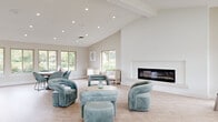 This thoughtfully designed single-floor space combines contemporary aesthetics with versatile functionality across several distinct areas. The expansive main lounge features vaulted, beam-accented ceilings, a sleek linear fireplace set within a crisp white surround, and abundant natural light streaming through multiple large windows that frame lush outdoor greenery. Light-toned herringbone-patterned flooring flows throughout, anchoring the space in warmth and elegance. Plush teal velvet seating arrangements create inviting conversation areas, while a dining setup with coordinating chairs offers a more structured gathering option. An adjoining game room provides generous open space with a billiards table, arcade game, a long built-in bar-height counter with seating, and continued access to natural light through panoramic windows. A private office area features a striking textured accent wall in soft wood-tone tones, a marble-topped white desk, and direct sightlines into the lounge. A dedicated workstation room supports administrative functions, and a fully equipped fitness center enclosed in floor-to-ceiling glass panels houses cardio machines, free weights, and mirrored walls. A quiet reading nook with a rattan chair and garden views completes the space.