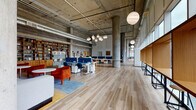 This expansive single-floor workspace blends industrial architecture with thoughtfully curated interiors to create a versatile and inspiring environment. Exposed concrete columns, soaring ceilings with visible ductwork, and polished or wood-plank flooring throughout establish a raw yet refined aesthetic. Floor-to-ceiling windows along one side of the space flood the interior with natural light while offering sweeping urban views of the surrounding cityscape.

The open-plan work area features adjustable sit-stand desks with blue privacy dividers, ideal for focused individual work or collaborative sessions. A floor-to-ceiling built-in bookcase, fully stocked with books and accessories, anchors a comfortable lounge zone furnished with vibrant orange velvet seating and colorful area rugs. Pendant globe light clusters add warm, designer-grade lighting throughout.

A standout lounge nook features a bold, botanical mural covering the full wall, complemented by a wraparound teal sectional sofa and warm wood coffee tables — a visually striking retreat within the workspace. The fully equipped kitchen includes teal cabinetry, teal tile backsplash, double wall ovens, a large central island with bar seating, and a polished concrete floor. An entry lobby with a circular teal sofa, built-in wooden shelving, and staircase access completes this thoughtfully designed, community-oriented workspace.