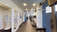 This fitness facility is designed to provide a comprehensive workout experience within a single-floor layout. The main exercise area stretches along an elongated floor plan, lined with a variety of cardio machines including treadmills, creating an organized and efficient use of space. Overhead, recessed and track lighting illuminates the room evenly, contributing to a bright and energizing atmosphere. The walls are finished in a crisp white and accented with bold blue tones, giving the space a clean, modern aesthetic. Durable rubber flooring covers the workout area, offering both safety and comfort underfoot. Strength training equipment, including a cable pulley machine, is positioned along the perimeter, maximizing the usable floor space for movement and exercise. Toward the entrance, a restroom is conveniently accessible, featuring white walls and practical fixtures. Informational signage is posted along the corridor walls, keeping members informed and engaged. The overall ambiance is professional and functional, with a straightforward design that prioritizes utility and ease of use for fitness enthusiasts of all levels.