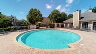 Located at 51 Northtown Dr in Jackson, Mississippi, this single-level outdoor amenity space is centered around a spacious, kidney-shaped swimming pool with clear turquoise water. The pool area is finished with attractive brick paver decking that surrounds the pool, providing ample room for lounging and relaxation. Poolside seating with chairs is conveniently arranged along the deck, offering a comfortable space for residents to unwind. A safety life ring and posted signage reflect a well-maintained environment. The pool is enclosed by a black iron security fence, ensuring a safe and private setting. A charming, low-profile accessory building with a pitched roof and large windows sits adjacent to the pool, providing additional utility or shelter for guests. Mature, leafy trees frame the perimeter of the space, offering natural shade and a tranquil, park-like ambiance. The open sky above and surrounding greenery create a bright, welcoming outdoor retreat. This well-appointed community amenity space combines practical design with an inviting atmosphere, making it an ideal destination for outdoor enjoyment in the heart of Jackson, MS.