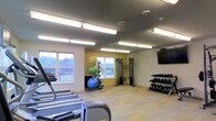 This fitness center offers a comprehensive workout environment designed to accommodate a variety of exercise routines. The space features light wood-toned flooring that contributes to a clean, modern aesthetic, while the gray ceiling with flush-mounted fluorescent lighting ensures the room is consistently and evenly illuminated throughout.

Large windows along the far wall invite natural light into the space and provide views of the surrounding greenery, creating an open and energizing atmosphere. An accent wall painted in a warm yellow tone adds a vibrant visual element to the room.

The equipment layout is well-organized and varied. Cardio machines, including treadmills, are positioned toward the front of the space. A cable resistance machine is centrally located, suitable for a wide range of strength training exercises. A dedicated free weight area along one wall features a neatly arranged dumbbell rack with a full range of weights. A medicine ball and stability ball storage rack occupies another corner, supporting functional and core training. A wall-mounted flat-screen television is positioned for easy viewing during workouts, adding to the overall convenience of the space.