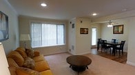 This two-floor space presents a clean, functional layout with a welcoming living area as a central feature. The living room is finished with light-toned carpeting and neutral cream walls, creating a warm and understated atmosphere. A large window fitted with vertical blinds allows natural light to fill the room, complementing the recessed ceiling lighting overhead. Crown molding adds a subtle architectural detail along the upper edges of the walls, lending a polished look to the space. Visible HVAC vents are integrated into both the walls and floors, ensuring climate control throughout. Adjacent to the living area, the dining space transitions to hardwood-style flooring, visible through an open passageway. A door with a window in the dining area provides an additional source of natural light and potential access to an outdoor area. The overall design is straightforward and practical, with a neutral color palette that offers versatility for various furnishing styles. The open sightline between the living and dining areas enhances the sense of connectivity and spaciousness across the main level of this two-floor property.