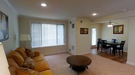 This two-floor space presents a clean, functional layout with a welcoming living area as a central feature. The living room is finished with light-toned carpeting and neutral cream walls, creating a warm and understated atmosphere. A large window fitted with vertical blinds allows natural light to fill the room, complementing the recessed ceiling lighting overhead. Crown molding adds a subtle architectural detail along the upper edges of the walls, lending a polished look to the space. Visible HVAC vents are integrated into both the walls and floors, ensuring climate control throughout. Adjacent to the living area, the dining space transitions to hardwood-style flooring, visible through an open passageway. A door with a window in the dining area provides an additional source of natural light and potential access to an outdoor area. The overall design is straightforward and practical, with a neutral color palette that offers versatility for various furnishing styles. The open sightline between the living and dining areas enhances the sense of connectivity and spaciousness across the main level of this two-floor property.