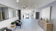 This single-floor space presents a sleek, contemporary lobby designed with both functionality and aesthetics in mind. The entryway is anchored by large-format light gray porcelain tile flooring that extends throughout, lending the space an open, polished feel. Recessed ceiling lighting, paired with a flush-mount HVAC unit set within a grid-panel ceiling, ensures the area is well-lit and climate-controlled year-round.

A built-in bank of gray metal mailboxes lines one wall, offering organized mail access for building occupants. Adjacent to the mailboxes, a warm wood-toned credenza with herringbone-patterned cabinet fronts provides a decorative accent, topped with a stylish ceramic vase. On the opposite side, a comfortable gray accent chair and a small side table create a welcoming seating nook, complemented by potted greenery that adds a touch of life and color to the neutral palette.

The lobby benefits from natural light streaming through the glass entry door, which opens directly to the exterior and provides views of the surrounding area. A frosted glass partition wall adjacent to the entry hints at an adjoining office or management space. Security is supported by visible surveillance cameras positioned at the ceiling. Overall, the lobby strikes a balance between modern design and practical building amenities.