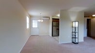 This single-floor space features an open layout that connects the main living area to the kitchen. The living area is finished with neutral-toned carpet and warm beige walls, creating a clean and inviting atmosphere. A front entry door with a multi-pane window allows natural light to filter into the space, complemented by a chandelier overhead that adds a modest decorative touch. A wall-mounted ductless air conditioning unit provides climate control directly within the main living area.

The kitchen is partially open to the living space, separated by a partial wall that defines the two areas without fully enclosing them. Dark wood cabinetry lines the kitchen, contrasting with lighter wall tones, and a stainless steel refrigerator is visible from the main living area. The kitchen features tile or hard flooring, distinct from the carpeted living space. Overall, the unit presents a straightforward, functional design with a neutral color palette throughout, offering a comfortable and practical living environment suitable for everyday use.