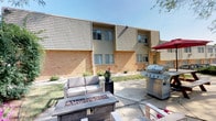 This building presents a classic brick-and-wood-siding exterior, lending it a well-established, solid character. The shared outdoor patio area is thoughtfully appointed with cushioned lounge seating, a rectangular fire pit table, and a large gas grill shaded by an umbrella — creating a welcoming space for outdoor gatherings and relaxation. Mature plantings and flower accents line the building's facade, adding a touch of greenery to the surroundings.

Inside, the common areas are defined by neutral-toned walls and carpeted flooring throughout. Wide hallways are decorated with framed artwork, contributing a warm and inviting atmosphere as residents move between spaces. A carpeted stairwell fitted with sturdy wood handrails provides access between levels, with clean, light-colored walls and fire safety equipment prominently placed for accessibility.

Upper-level corridors feature the same consistent carpeting and wood-trimmed doorways, with sloped ceiling details that hint at the building's architectural character. Natural light filters in at corridor ends through exterior-facing windows, keeping the passageways bright and airy. An on-site fitness room, visible through a wood-framed glass door, offers residents convenient access to exercise equipment. Overall, the building maintains a functional, comfortable, and well-kept environment throughout its shared spaces.