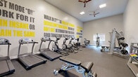 Located in Mill Creek, WA, this fitness center is a comprehensively outfitted facility designed to support a full range of workout routines across multiple distinct areas. The main cardio room features high ceilings, rubber flooring, and a striking accent wall adorned with bold motivational typography in gray and yellow, creating an energizing atmosphere. This area houses a row of treadmills and elliptical machines, complemented by ceiling fans that promote airflow throughout the space. A dedicated strength training zone provides rubber-floored workout space with a multi-function cable machine, adjustable bench, and a tiered dumbbell rack offering a range of weights. An adjoining room with rich hardwood flooring serves as a functional training and studio area, equipped with medicine ball racks, stability balls, and a large wall-mounted mirror ideal for form-checking during exercise. A bright spin studio features polished hardwood floors, multiple stationary cycling bikes, large windows that invite abundant natural light and views of surrounding greenery, and a ceiling fan overhead. Additional amenities include a wall-mounted display screen, a water dispenser, and French door access to the outdoors. Together, these spaces form a thoughtfully organized fitness facility suited for cardio, strength, and flexibility training.