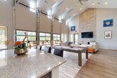 Located at 2523 Avenue M in Huntsville, Texas, this single-story community clubhouse offers a generously sized, open-concept gathering space designed for both social and functional use. The centerpiece of the facility is a dramatic great room defined by soaring vaulted ceilings with exposed angular rooflines, large industrial-style roll-up windows that flood the interior with natural light, and wide-plank hardwood-style flooring throughout. A striking floor-to-ceiling stacked stone accent wall anchors the lounge area, which features contemporary white seating and a wall-mounted flat-screen television. A full billiards table occupies the heart of the room, complemented by a nearby kitchenette with granite countertops, dark cabinetry, and bar seating—ideal for entertaining. The grand entrance foyer makes a bold first impression with soaring ceilings, curated artwork, a vibrant living green wall with a neon sign, and an abundance of open floor space. Two dedicated conference rooms provide a more focused environment, each equipped with a long work table, mixed seating, a whiteboard, and large windows overlooking the surrounding green landscape. A hallway connects the various zones, ensuring smooth circulation throughout the facility. The overall ambiance is modern, bright, and inviting.