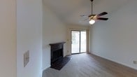 This single-floor space centers around a bright and open living area defined by its vaulted ceiling, which adds a sense of height and airiness to the room. A ceiling fan with a light fixture is mounted at the peak, providing both ventilation and ambient lighting. The living area is anchored by a tile-surround fireplace with a dark mantel, serving as a focal point along one wall. The hearth extends onto a tiled floor section that transitions into wall-to-wall carpeting throughout the rest of the room. Natural light enters generously through a sliding glass door that opens to an outdoor deck, offering views of the surrounding exterior. The neutral wall tones and clean lines throughout the space contribute to a simple, functional aesthetic. The combination of the vaulted ceiling, fireplace, and outdoor access creates a comfortable and versatile living environment suited to a range of uses.