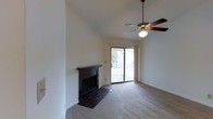 This single-floor space centers around a bright and open living area defined by its vaulted ceiling, which adds a sense of height and airiness to the room. A ceiling fan with a light fixture is mounted at the peak, providing both ventilation and ambient lighting. The living area is anchored by a tile-surround fireplace with a dark mantel, serving as a focal point along one wall. The hearth extends onto a tiled floor section that transitions into wall-to-wall carpeting throughout the rest of the room. Natural light enters generously through a sliding glass door that opens to an outdoor deck, offering views of the surrounding exterior. The neutral wall tones and clean lines throughout the space contribute to a simple, functional aesthetic. The combination of the vaulted ceiling, fireplace, and outdoor access creates a comfortable and versatile living environment suited to a range of uses.