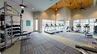 Located at 3 Woodcock Rd in Savannah, Georgia, this single-floor fitness center offers a well-appointed and thoughtfully designed workout facility. The main exercise area is distinguished by soaring vaulted ceilings clad in warm natural wood, exposed ductwork, and ceiling fans that lend an airy, industrial-chic aesthetic. Large windows line the walls, flooding the space with abundant natural light and providing views of the surrounding outdoor environment. The main floor is finished with patterned carpet tiles and houses a robust selection of cardio machines, including multiple treadmills, ellipticals, and a rowing machine, alongside a cable and functional trainer system and an adjustable weight bench. A dedicated strength and functional training zone features a multi-tier storage rack stocked with kettlebells, medicine balls, stability balls, and battle ropes, offering a comprehensive free-weight and conditioning experience. A separate room with hardwood flooring and sage-green walls serves as a cycling and spin studio, brightened by large corner windows with views of lush greenery. This room also contains a dumbbell rack and additional bikes, providing a quieter, focused training environment. Together, the two areas create a versatile and fully equipped fitness facility suited for a wide range of training styles and fitness goals.