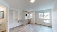 This well-designed single-floor studio offers a clean, modern aesthetic built around an efficient open-plan layout. Light wood-look luxury vinyl plank flooring runs throughout the entire unit, contributing to a bright and cohesive feel. The main living area is open and versatile, bathed in natural light from large windows that offer views of surrounding greenery.

The kitchenette is thoughtfully integrated into the space, featuring light wood-grain cabinetry, a compact cooktop, a single-basin sink, a stainless steel refrigerator, and an over-the-counter microwave. Adjacent to the kitchen, a built-in shelving unit with open cubbies and a fold-down wall-mounted desk provides flexible storage and workspace solutions without consuming additional floor space.

The bathroom is neatly appointed with white walls, wood-look flooring, a wall-mounted sink, a framed mirror, and a standard toilet, maintaining the clean and minimal design language found throughout the unit. Flush-mounted circular ceiling lights provide consistent, even illumination across all areas. The entry features a secure door with modern hardware, and the overall palette of white walls and natural wood tones creates a fresh, contemporary atmosphere well-suited for efficient, comfortable living.
