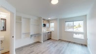 This well-designed single-floor studio offers a clean, modern aesthetic built around an efficient open-plan layout. Light wood-look luxury vinyl plank flooring runs throughout the entire unit, contributing to a bright and cohesive feel. The main living area is open and versatile, bathed in natural light from large windows that offer views of surrounding greenery.

The kitchenette is thoughtfully integrated into the space, featuring light wood-grain cabinetry, a compact cooktop, a single-basin sink, a stainless steel refrigerator, and an over-the-counter microwave. Adjacent to the kitchen, a built-in shelving unit with open cubbies and a fold-down wall-mounted desk provides flexible storage and workspace solutions without consuming additional floor space.

The bathroom is neatly appointed with white walls, wood-look flooring, a wall-mounted sink, a framed mirror, and a standard toilet, maintaining the clean and minimal design language found throughout the unit. Flush-mounted circular ceiling lights provide consistent, even illumination across all areas. The entry features a secure door with modern hardware, and the overall palette of white walls and natural wood tones creates a fresh, contemporary atmosphere well-suited for efficient, comfortable living.