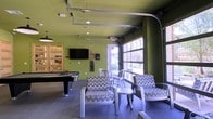 This energetic amenity lounge is designed to offer a dynamic and social atmosphere for its users. The space is defined by its bold, lime-green walls that create a lively and inviting ambiance, complemented by tile flooring throughout. At the heart of the room, a pool table serves as a central entertainment feature, positioned alongside a wall-mounted flat-screen television, making it an ideal gathering spot for recreation and relaxation. A rack of billiard cues is conveniently mounted nearby, adding to the room's game-ready setup. The lounge area is furnished with chevron-patterned accent chairs and small side tables, creating a comfortable seating arrangement that encourages conversation and socializing. One of the most distinctive architectural features is the large, industrial-style glass garage door that opens to bring the outdoors in, allowing an abundance of natural light to pour into the space and offering views of the surrounding exterior. Exposed overhead tracks for the garage door add an industrial design touch to the ceiling. An adjacent windowed room, accessible through a slatted wood-and-glass door, provides a secondary space that adds functional versatility to the overall layout. The combination of bold color, open design, and varied amenities makes this a well-appointed communal space.