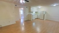 This single-floor space features an open-concept design that integrates the living area and kitchen into one cohesive layout. The flooring throughout is a warm, light-toned hardwood-style material that enhances the brightness of the space and creates a clean, uniform look.

The living area is spacious and well-lit, equipped with a ceiling fan with a light fixture that provides both airflow and overhead lighting. Visible HVAC vents in the ceiling suggest a central air system throughout the unit.

The kitchen is positioned at the rear of the open area and features white cabinetry, white appliances including a gas range, dishwasher, and a full-sized refrigerator. A sink is centrally located along the counter, and under-cabinet lighting adds functional illumination to the workspace. The kitchen maintains a clean, practical aesthetic consistent with the rest of the space.

A hallway visible from the main living area leads to additional rooms, with natural light filtering through a window at the far end, contributing to the overall brightness of the interior. The neutral, cream-toned walls throughout keep the space feeling open and versatile.