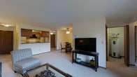 This single-floor residence offers an open and functional layout that seamlessly connects the main living areas. The living room is carpeted and comfortably sized, featuring a ceiling fan for year-round comfort and ample space for seating and entertainment. A media console with a large flat-screen television anchors the space, while a glass-topped coffee table and patterned armchair add a casual, welcoming feel. The living area flows naturally into a dedicated dining space, which accommodates a small table and chairs beneath another ceiling fan, creating a defined yet open area for everyday meals. Adjacent to the dining space, the kitchen is appointed with warm wood-toned cabinetry, a white refrigerator, and a built-in microwave above the range. A peninsula counter separates the kitchen from the living and dining areas, providing both additional prep space and a casual breakfast bar option. Light-toned carpeting runs throughout the main living areas, complemented by white walls that keep the space feeling bright and open. Neutral tones and practical finishes give the home a clean, move-in ready character. A hallway visible from the main area suggests additional private spaces beyond the main living zone.